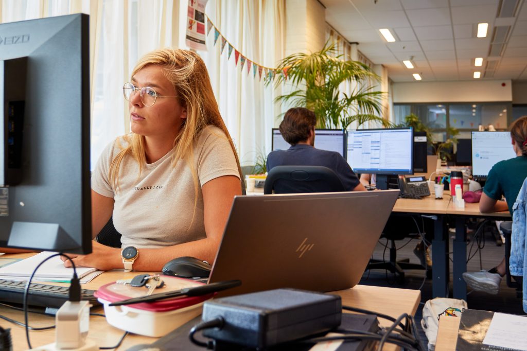 Vrouw met bril werkt op computer in modern kantoor met grote ramen en planten; anderen werken ook achter hun schermen.