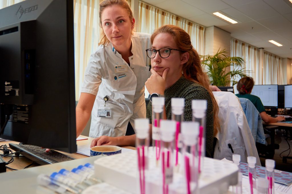 Twee vrouwen werken aan een computer in een lab, met reageerbuizen met roze vloeistof op de voorgrond.
