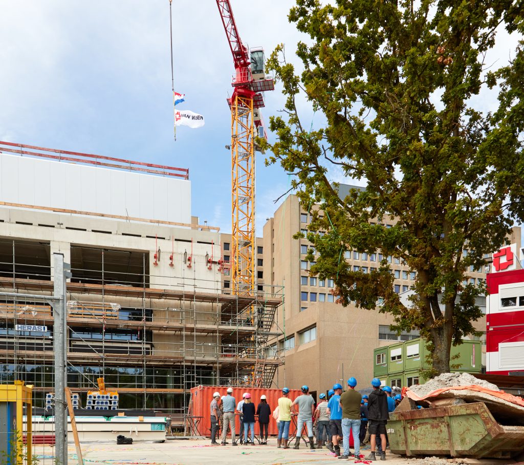Mensen met helmen op de bouwplaats bij hoogste punt gebouw R kijken naar een kraan. Stuurlui, gebouwen en een boom zichtbaar.
