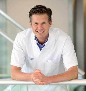 Oncologisch chirurg Misha Luyer in witte jas leunt op reling en glimlacht. Achtergrond: lichtgekleurde muren en metalen balustrades.