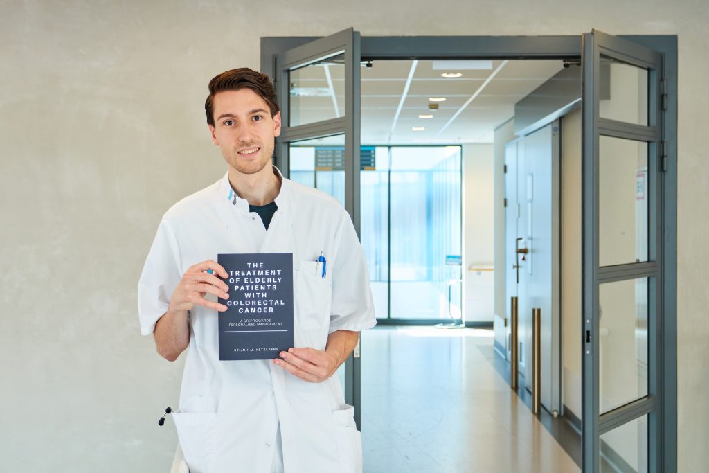 Persoon in witte labjas staat in gang met boek over darmkanker bij ouderen. Achter hem open deuren naar heldere bende.