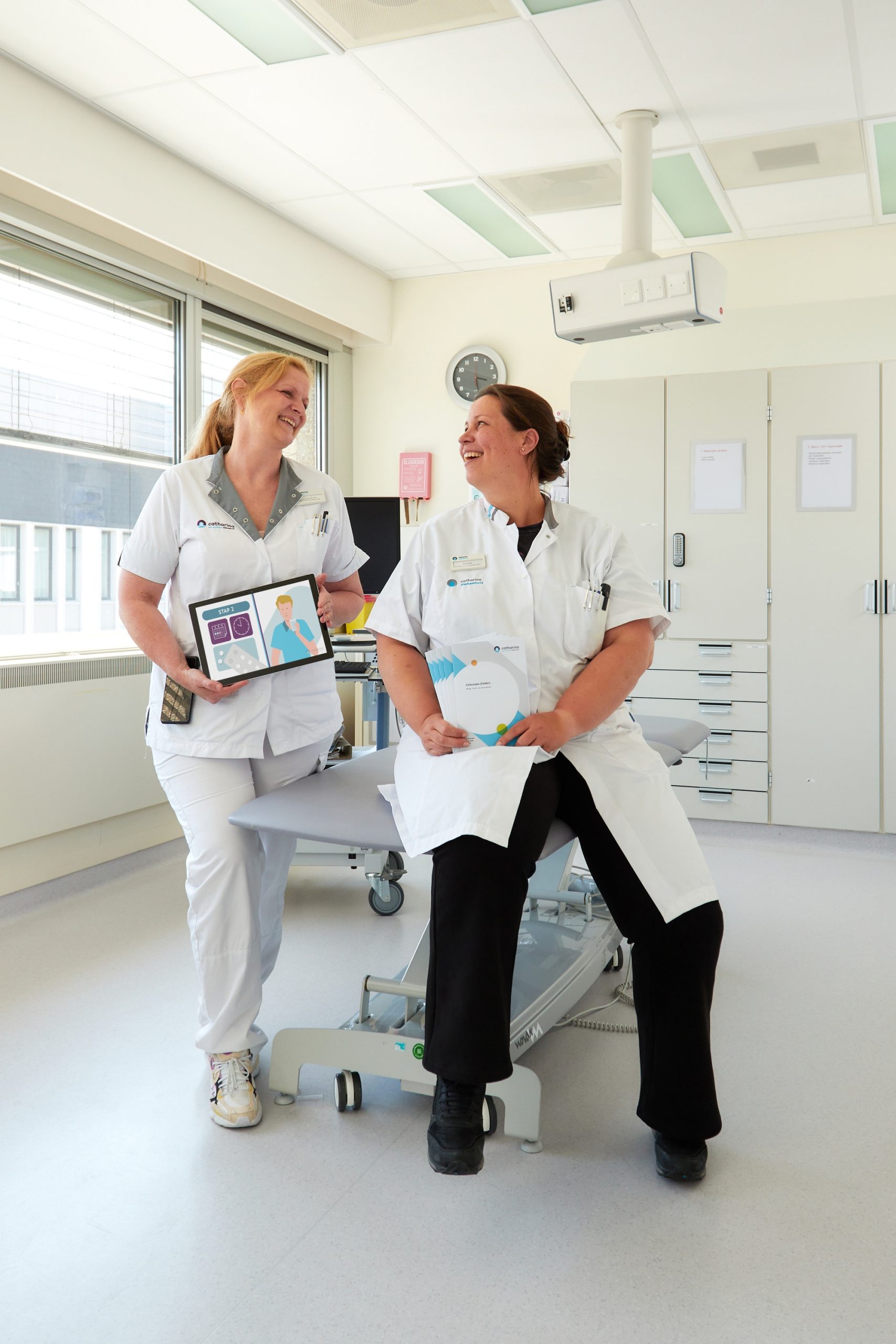 Twee zorgverleners in witte uniformen bespreken coloscopievoorlichting met tablet en kaart in een heldere behandelkamer.