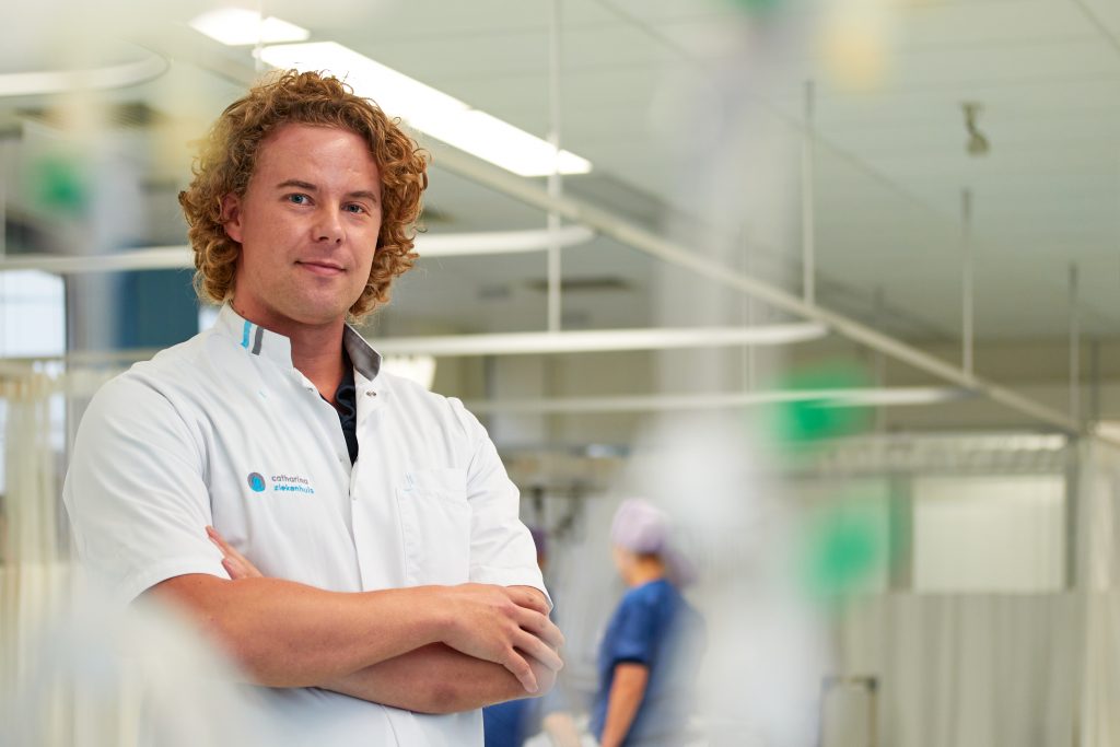 Zorgverlener met krullend haar, armen over elkaar, in kliniek bij hartpatiënten. Persoon in scrubs op de achtergrond.