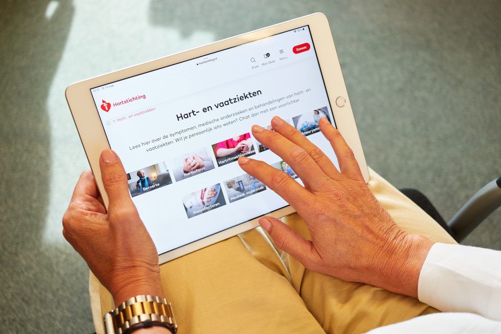 Persoon met goudkleurig horloge en beige broek houdt tablet vast met Nederlandstalige hart- en vaatwebsite voor patiënten.