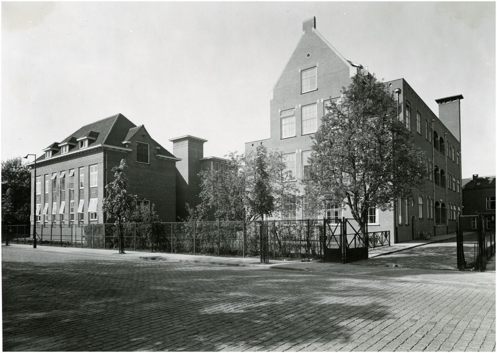 Zwart-witfoto van twee bakstenen gebouwen met een binnenplaats, bomen en hek; kinderkopstraat, geen mensen of voertuigen.