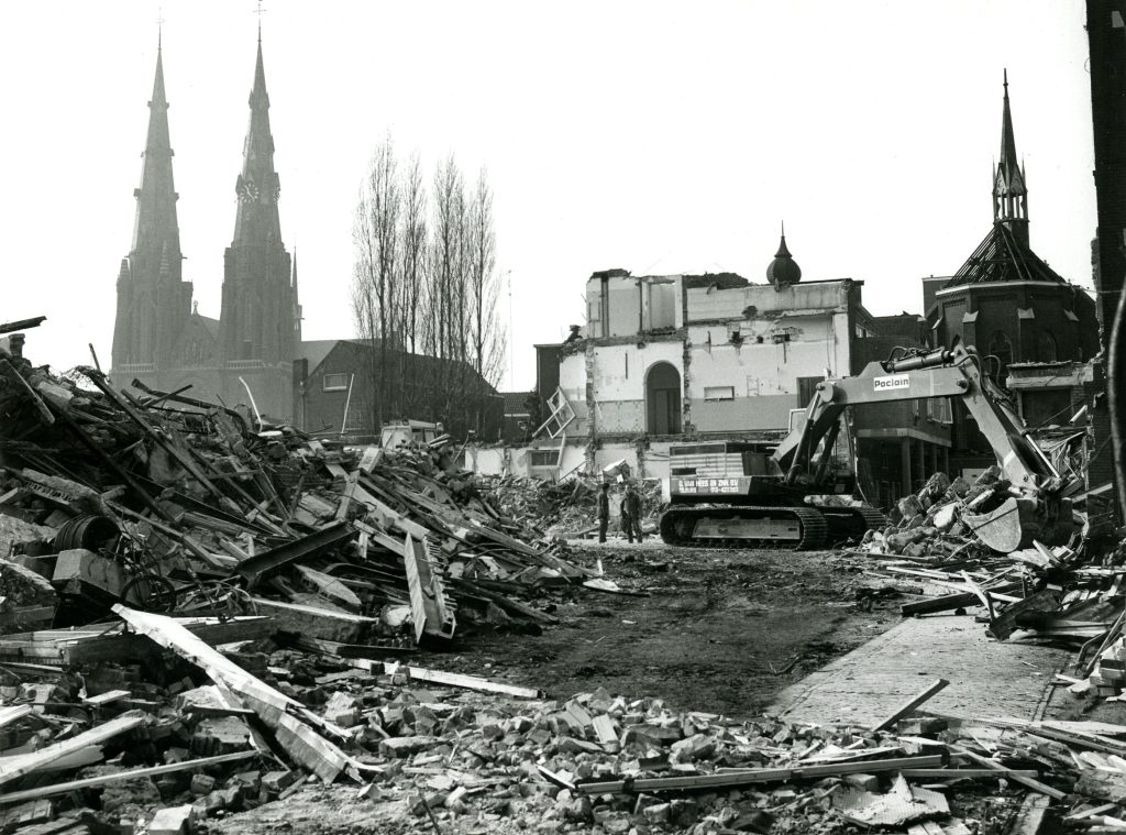 Een graafmachine ruimt puin tussen gesloopte gebouwen, met kerktorens en het Catharina Ziekenhuis op de achtergrond in de mist.