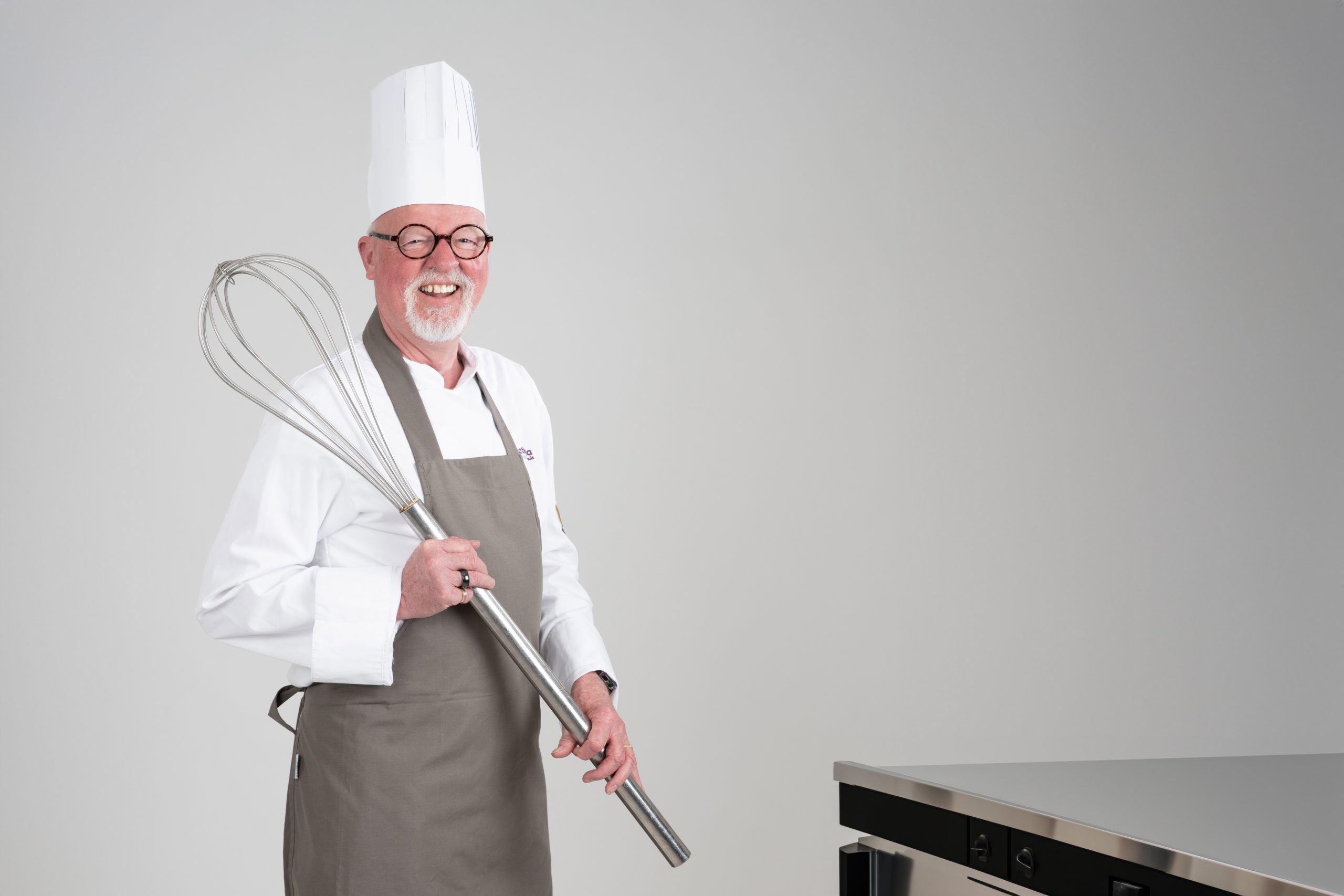Een lachende chef in wit uniform met grote garde bij metalen aanrecht, viert vijftigjarig jubileum van Catharina Ziekenhuis.