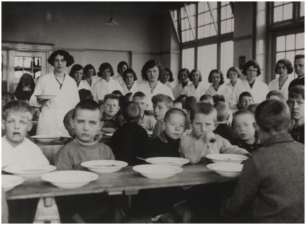 Kinderen zitten aan lange tafels met lege kommen; vrouwen in witte uniformen staan op de achtergrond in het Catharina Ziekenhuis.