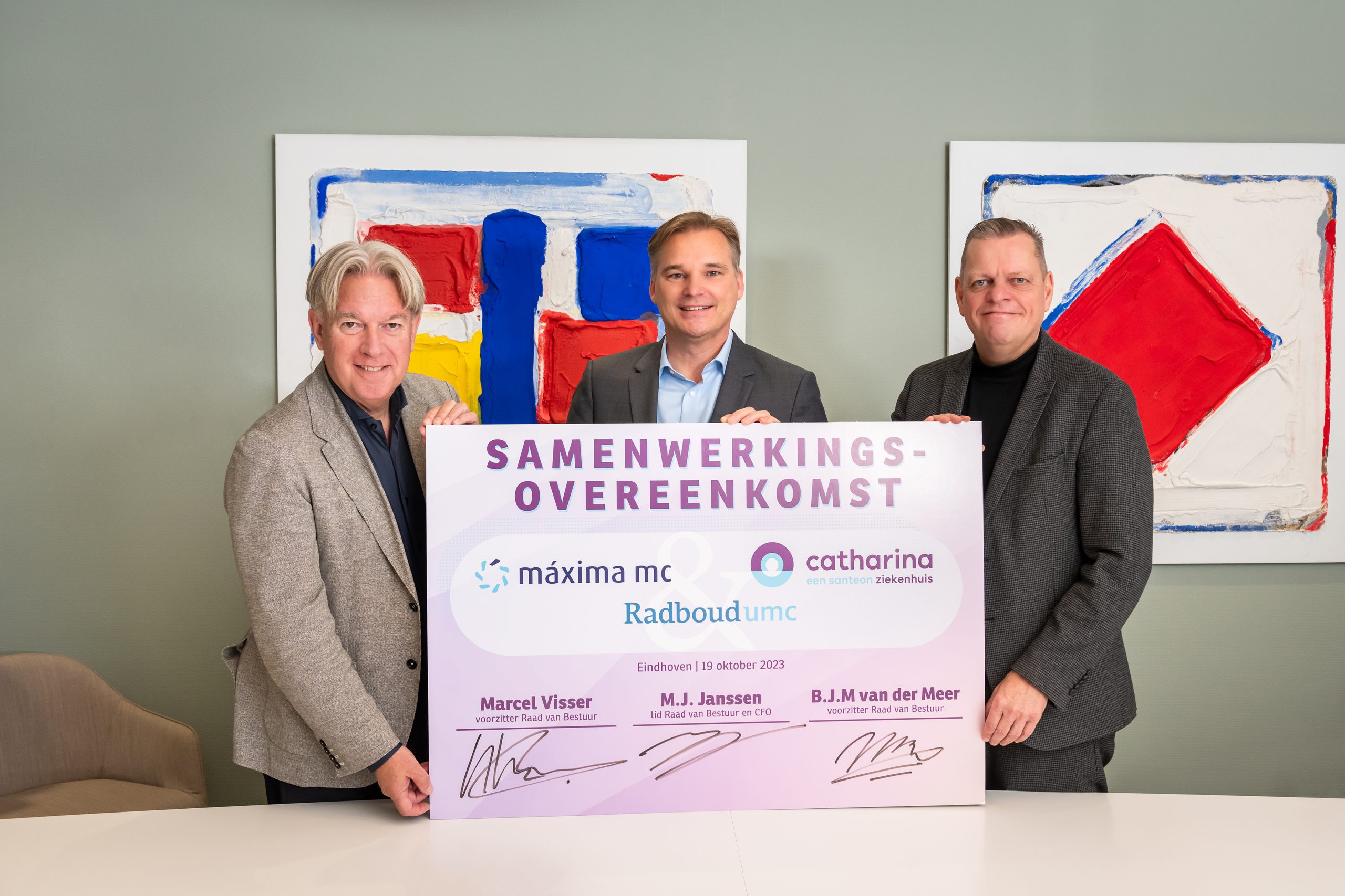 Drie mannen in pak achter bord over samenwerking Máxima MC, Catharina Ziekenhuis en Radboudumc. Abstract kunstwerk achter hen.