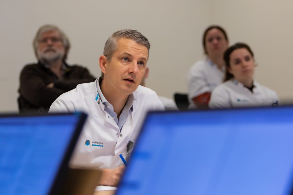 Man in witte labjas aan tafel luistert bij vergadering over endeldarmkanker; drie collega's en computerschermen op achtergrond.