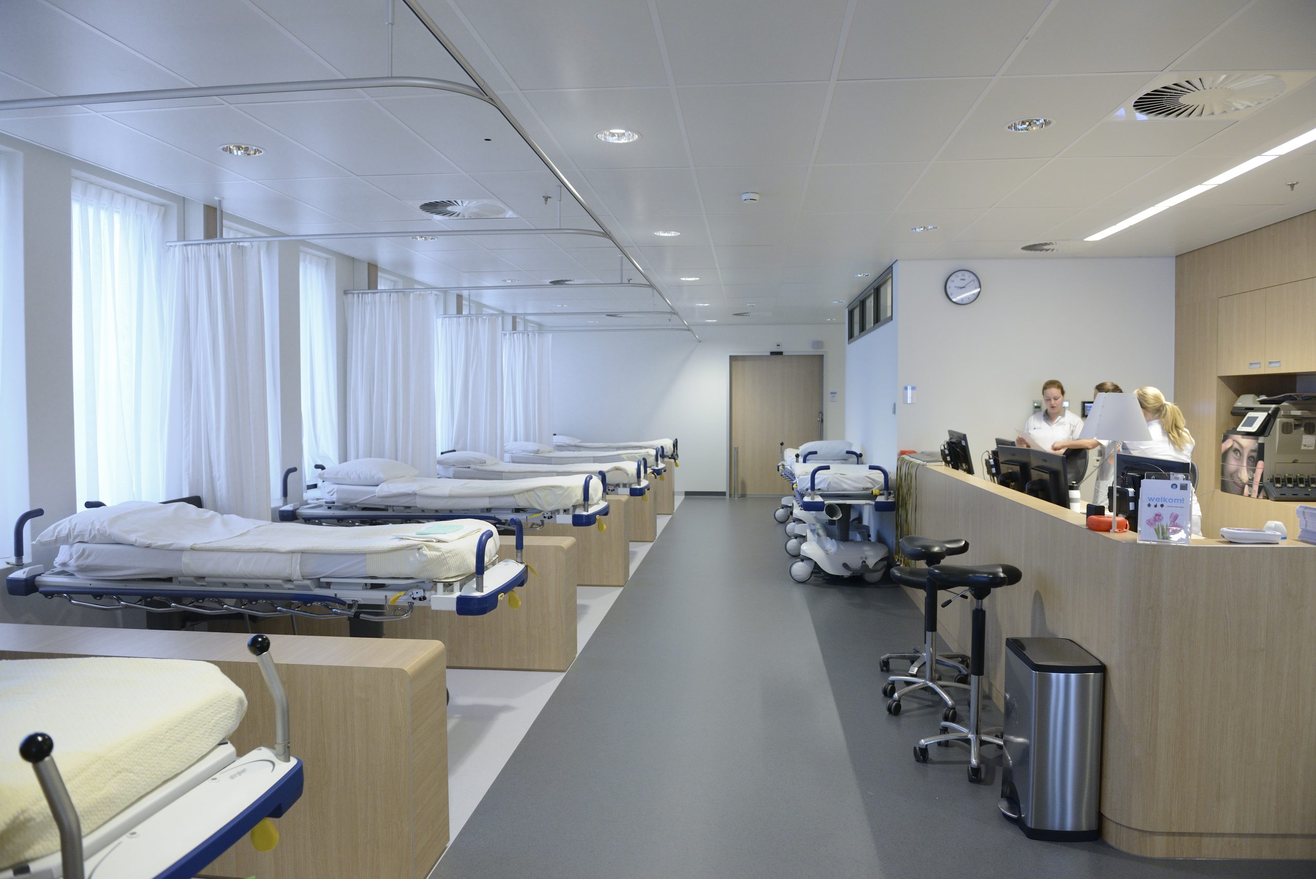 Een ziekenhuiszaal met lege bedden gescheiden door gordijnen, een verpleegpost en medische apparatuur langs de muren.