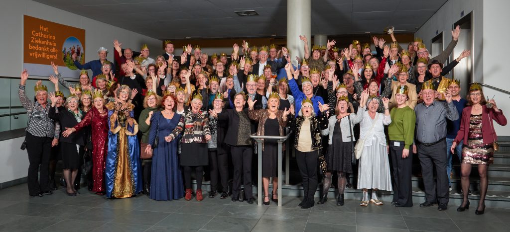 Grote groep mensen in feestelijke kleding op een val, lachend met opgestoken armen. Bord vermeldt 35 jaar vrijwilligers.