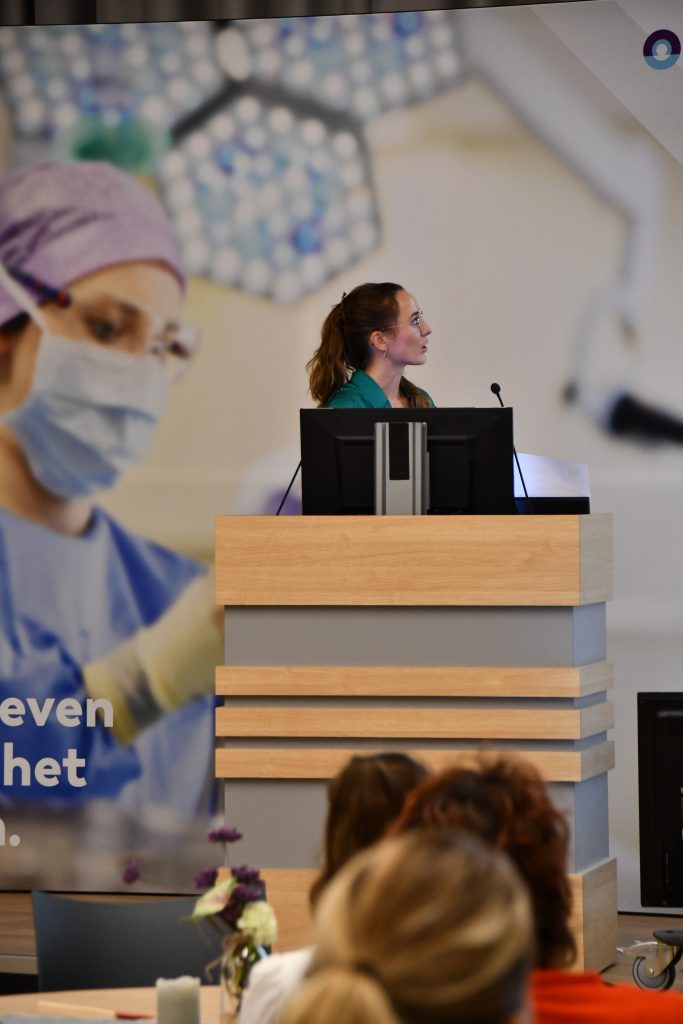 Vrouw presenteert over chirurgische oncologie bij een lessenaar met twee schermen; publiek en medische poster op achtergrond.