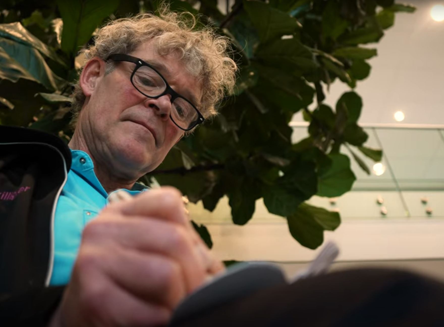 Pim Beishuizen schrijft in een notitieboek. Hij draagt een blauw shirt en donker colbert, voor grote groene planten.