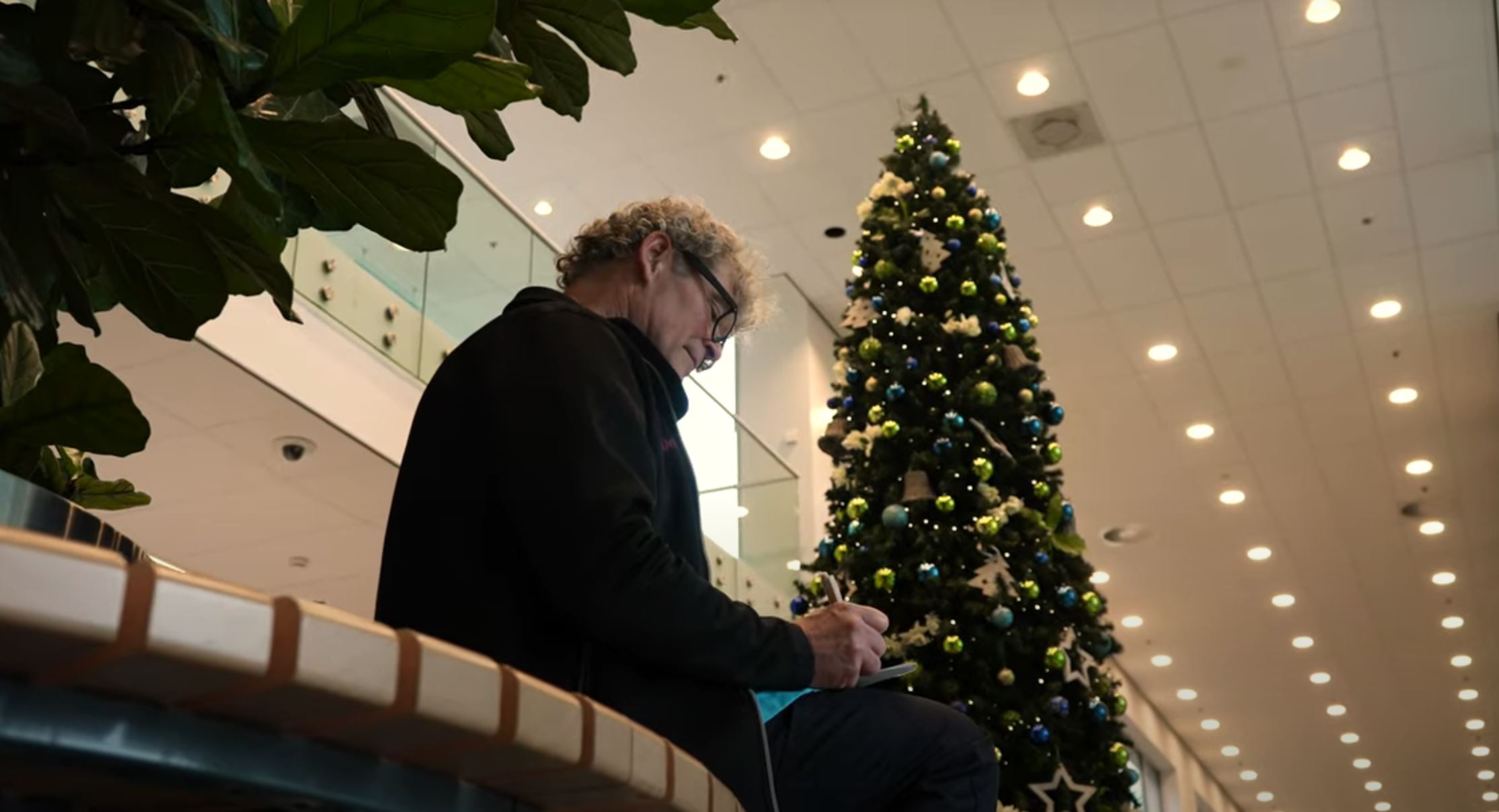 Persoon met grijs krullend haar schrijft op een notitieblok in moderne ruimte met hoge kerstboom met blauwe, groene en witte ballen.