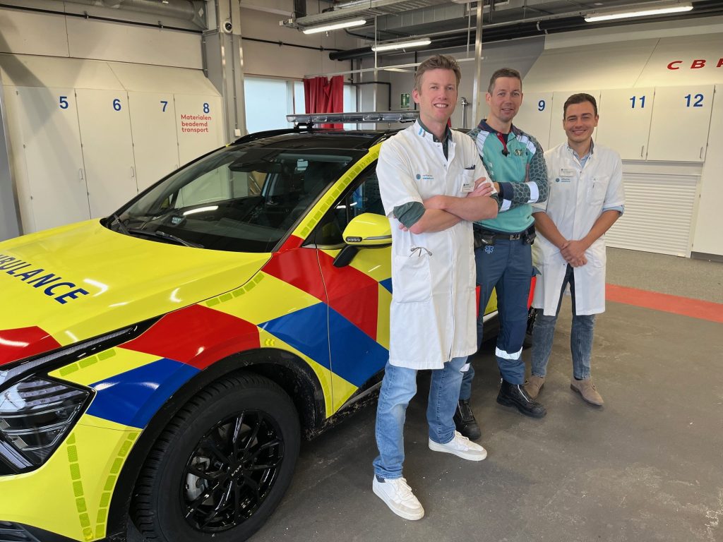 Drie zorgverleners bij een ambulance in een garage; twee in witte jassen, één in groen uniform. Genummerde deuren achter hen.