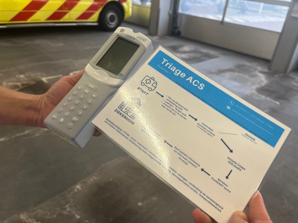 Persoon houdt een medisch apparaat en een gelamineerd Triage ACS-schema vast; op de achtergrond staat een ambulance.