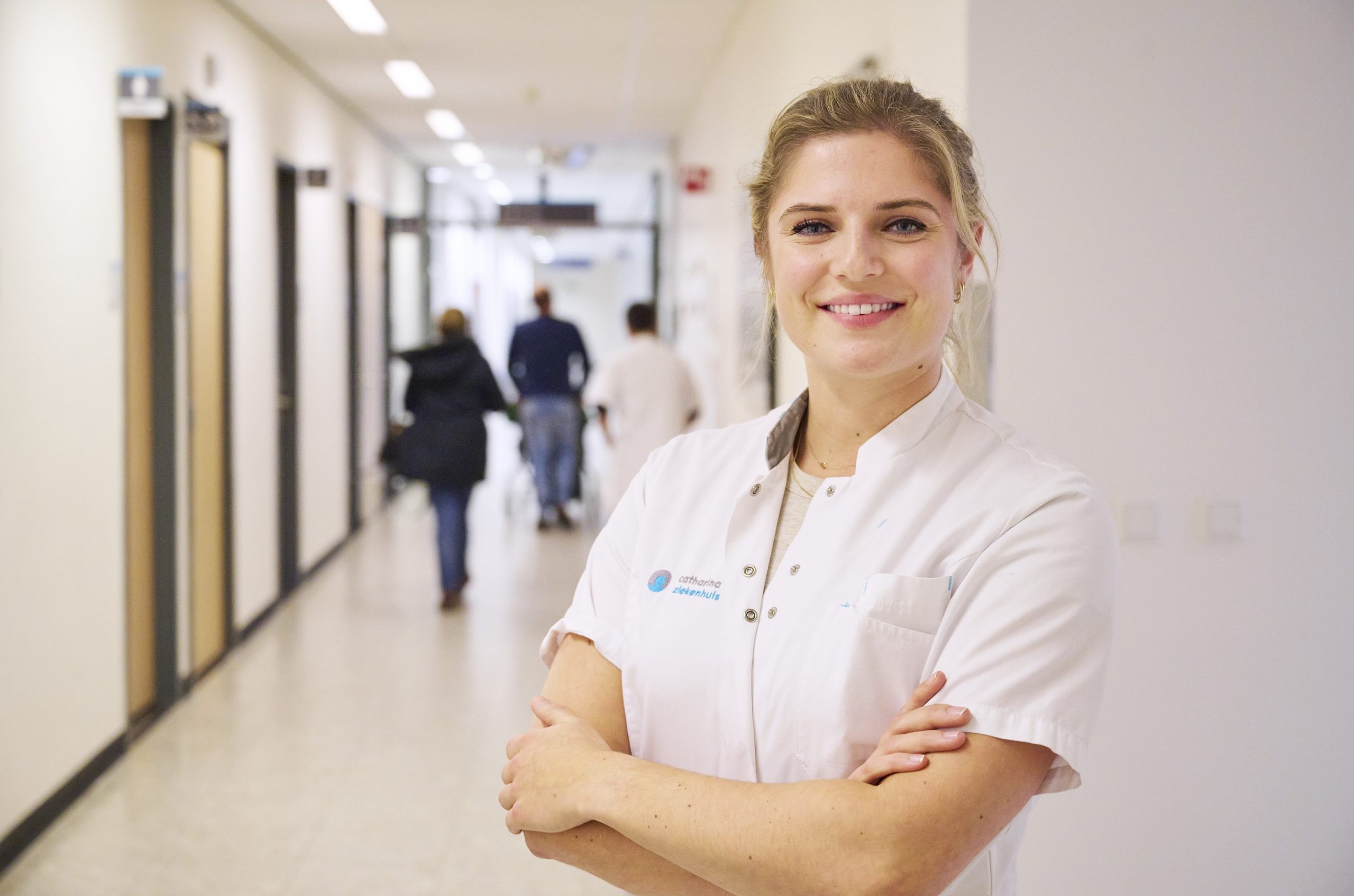 Vrouw in witte doktersjas staat met gekruiste armen en glimlacht in heldere zorggang, mensen lopen op achtergrond.