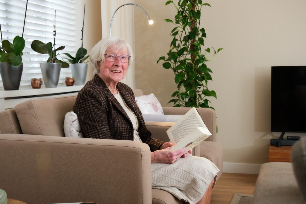Oudere vrouw met wit haar en bril zit op beige fauteuil met open boek. Op de achtergrond planten, lamp en tv in zorgwoning.