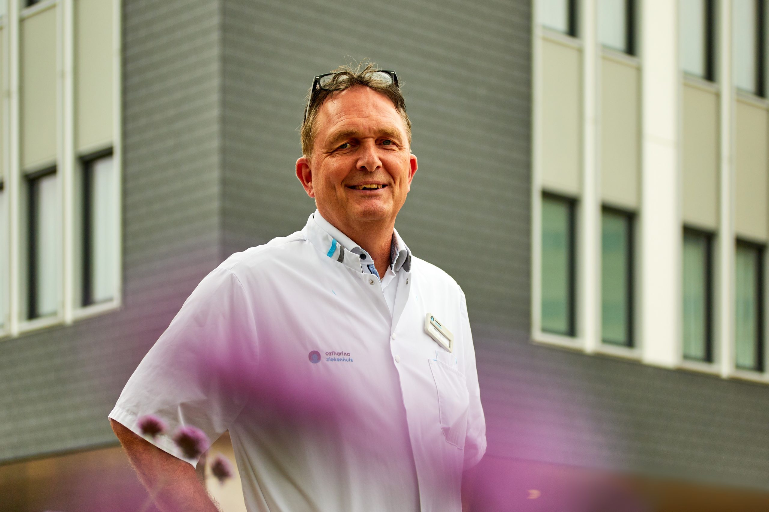 Man in witte doktersjas staat lachend voor modern gebouw; vage paarse bloemen op voorgrond, verwijzing naar thuismonitoring.