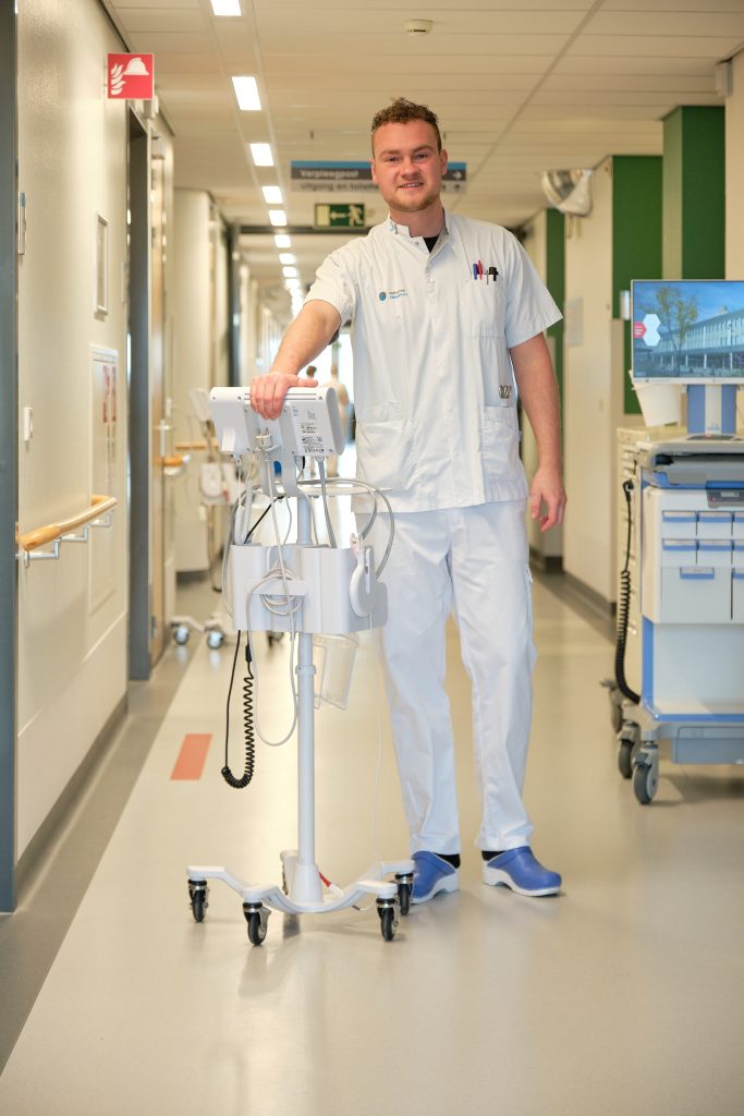Verpleegkundige Jelle van de Boomen in witte kleding staat in een heldere ziekenhuisgang en roest met één hand op een medisch apparaat op wielen.