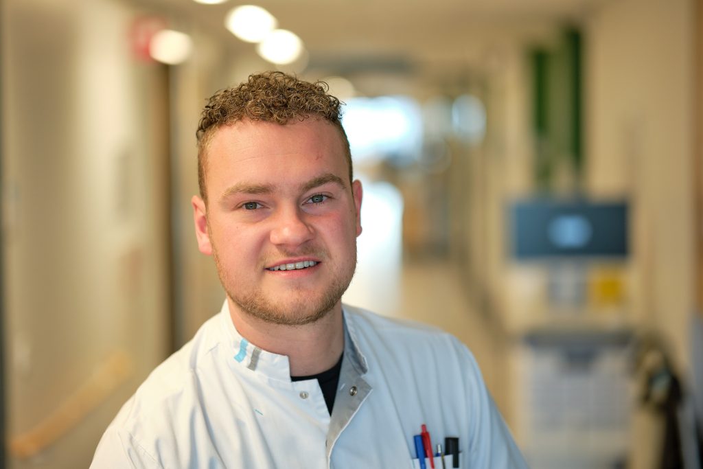 Verpleegkundige Jelle van de Boomen in witte jas staat lachend in een heldere gang met vage medische apparatuur.