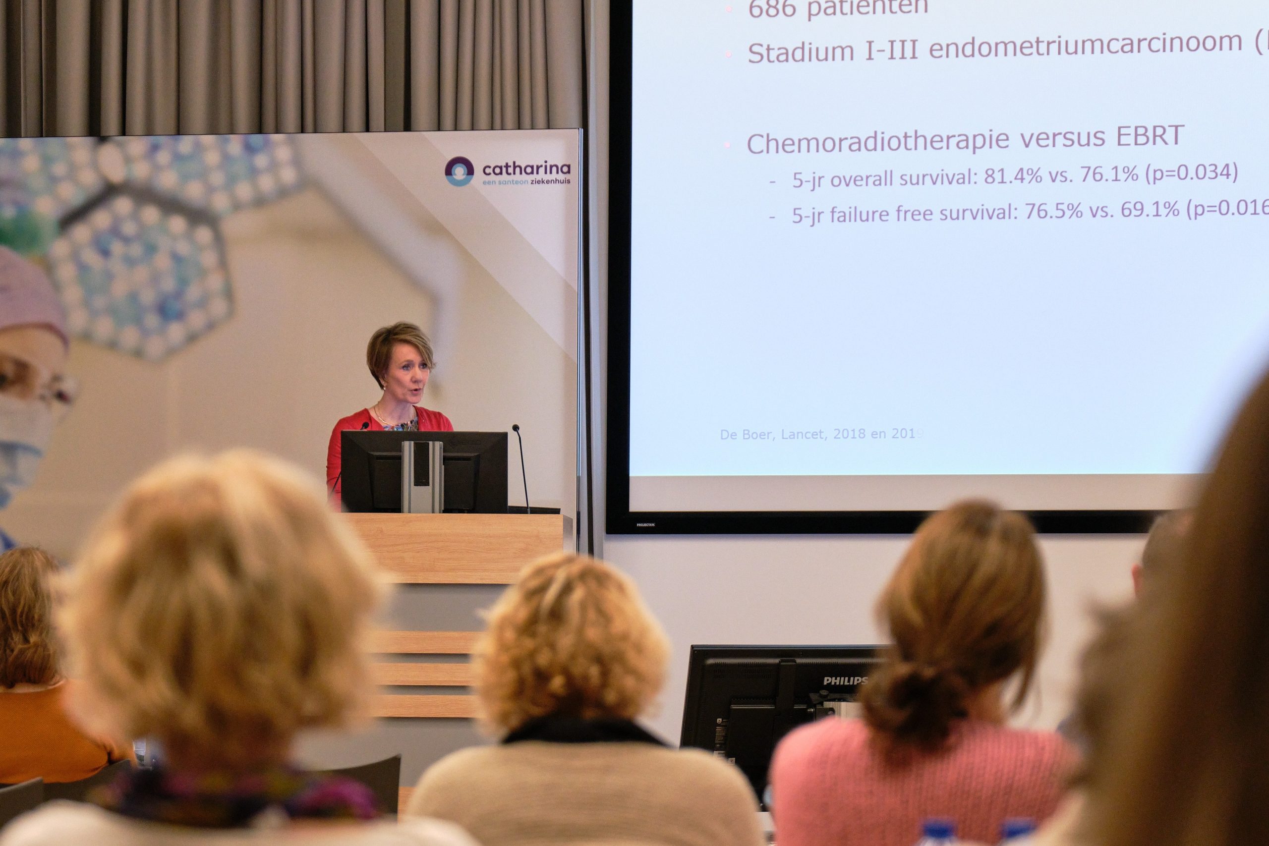 Vrouw presenteert zich bij een katheder; scherm toont dia's over endometriumcarcinoom, radiotherapie en nieuwe behandelingen.
