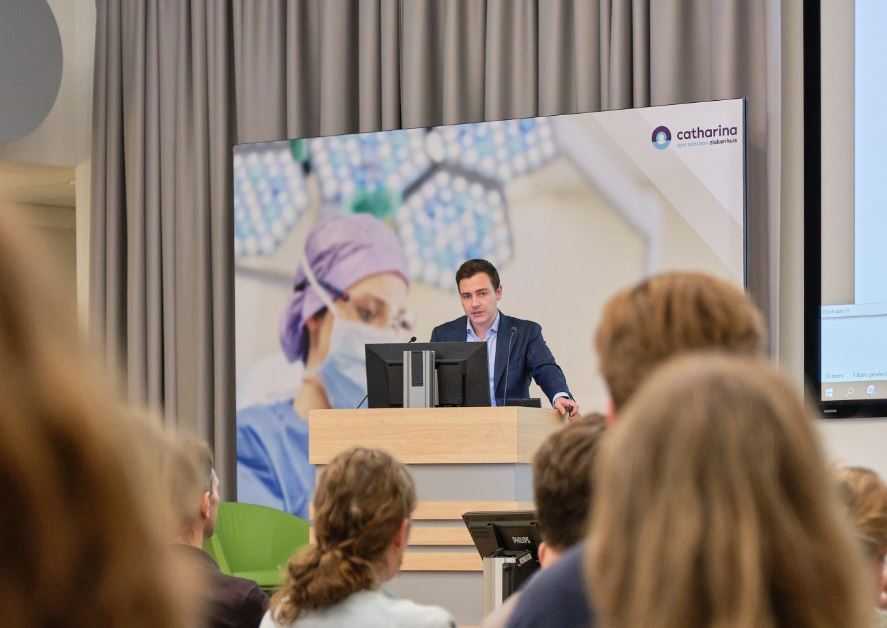 Man in pak geeft presentatie over hartzorg bij een katheder; op achtergrond foto van zorgverlener en Catharina Ziekenhuis logo.