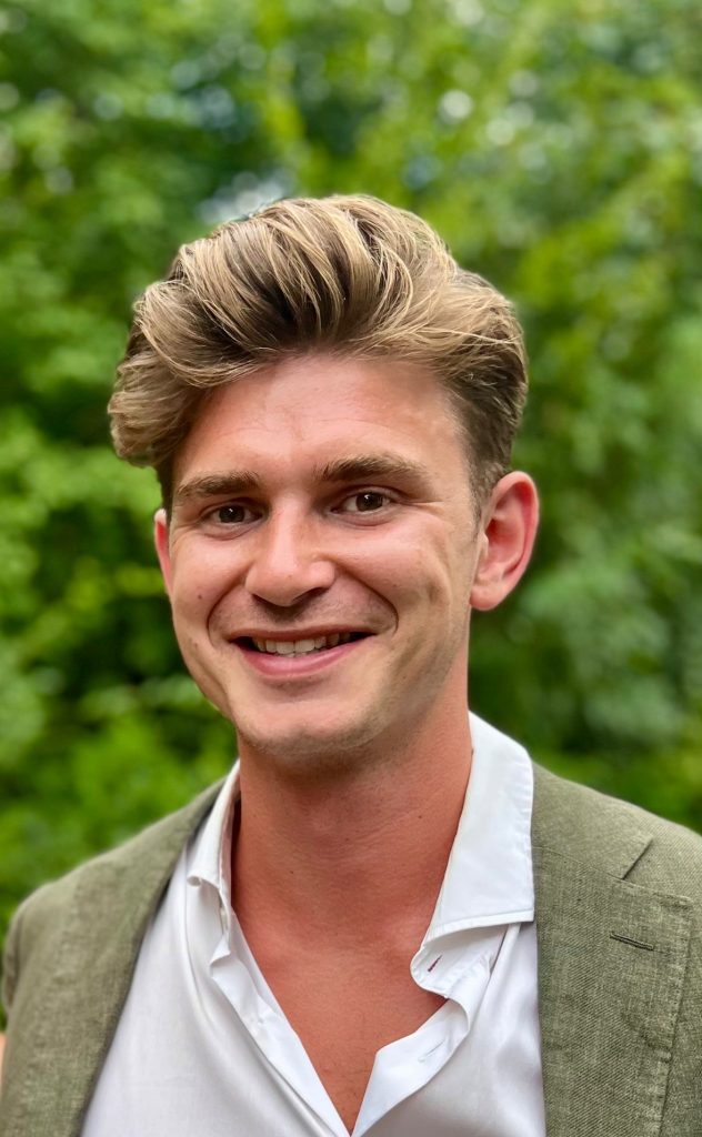 Man met kort, lichtbruin haar lacht naar de camera in witte blouse en olijfgroene blazer, voor groene bomen.