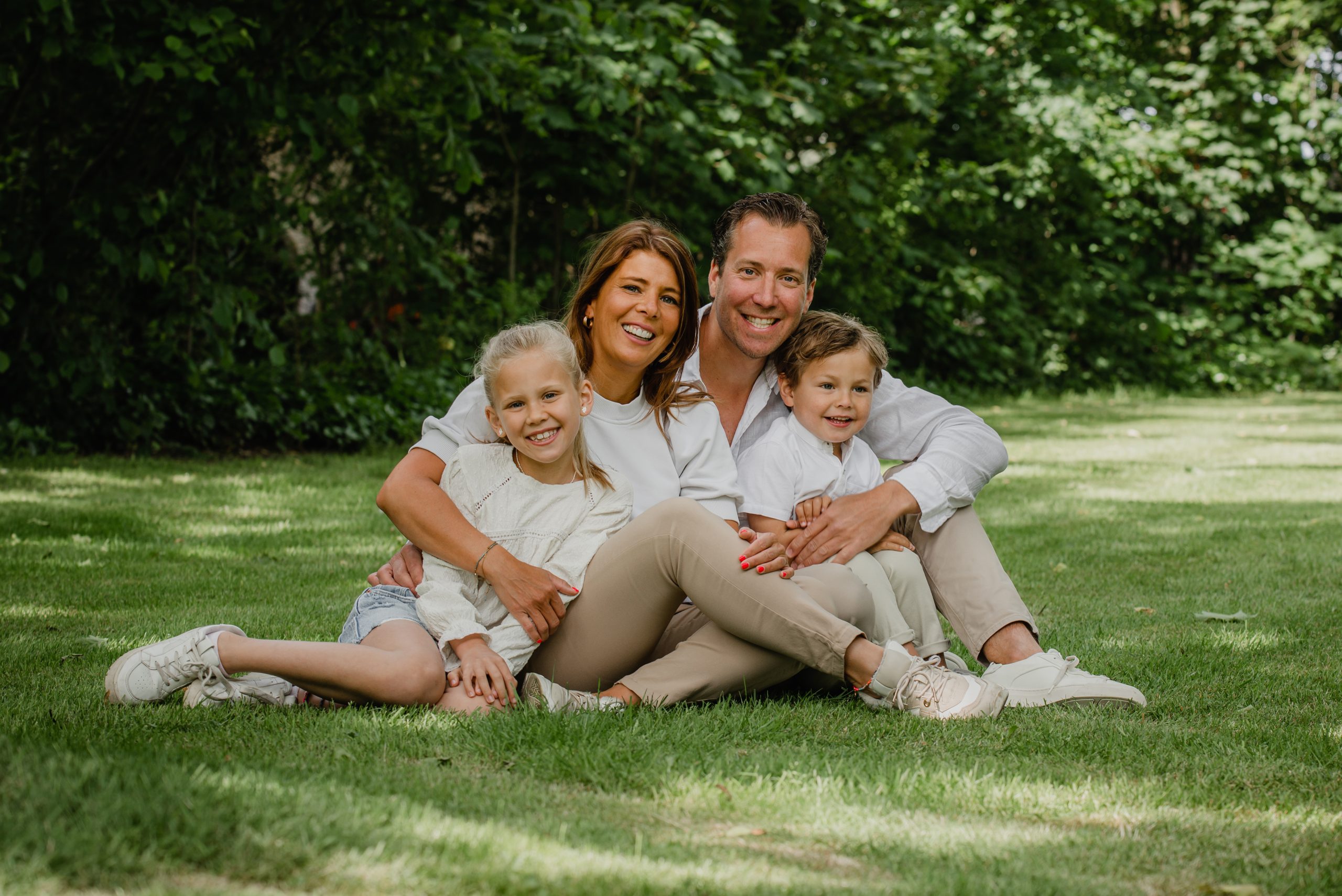 Man en vrouw zitten met twee kinderen op het gras buiten, lachend naar de camera. Op de achtergrond staan bomen en groen.