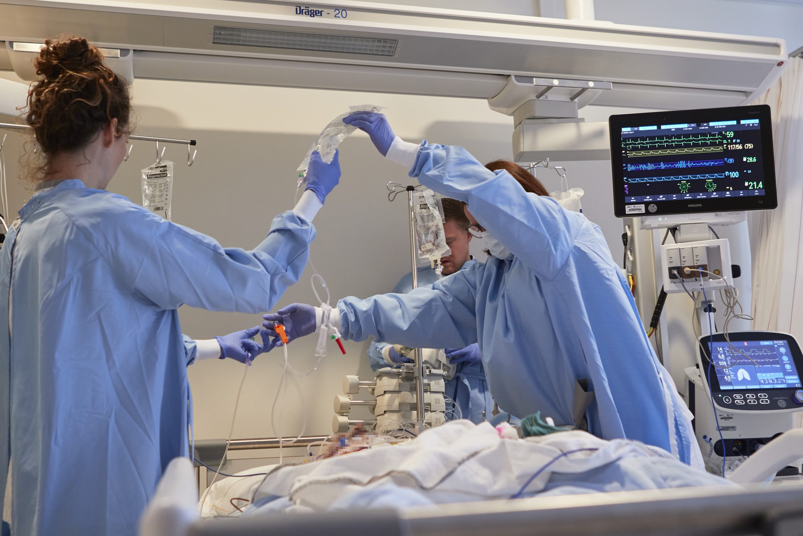 Drie zorgverleners in blauwe schorten zorgen voor een IC-patiënt in het Catharina Ziekenhuis, omringd door medische apparatuur.