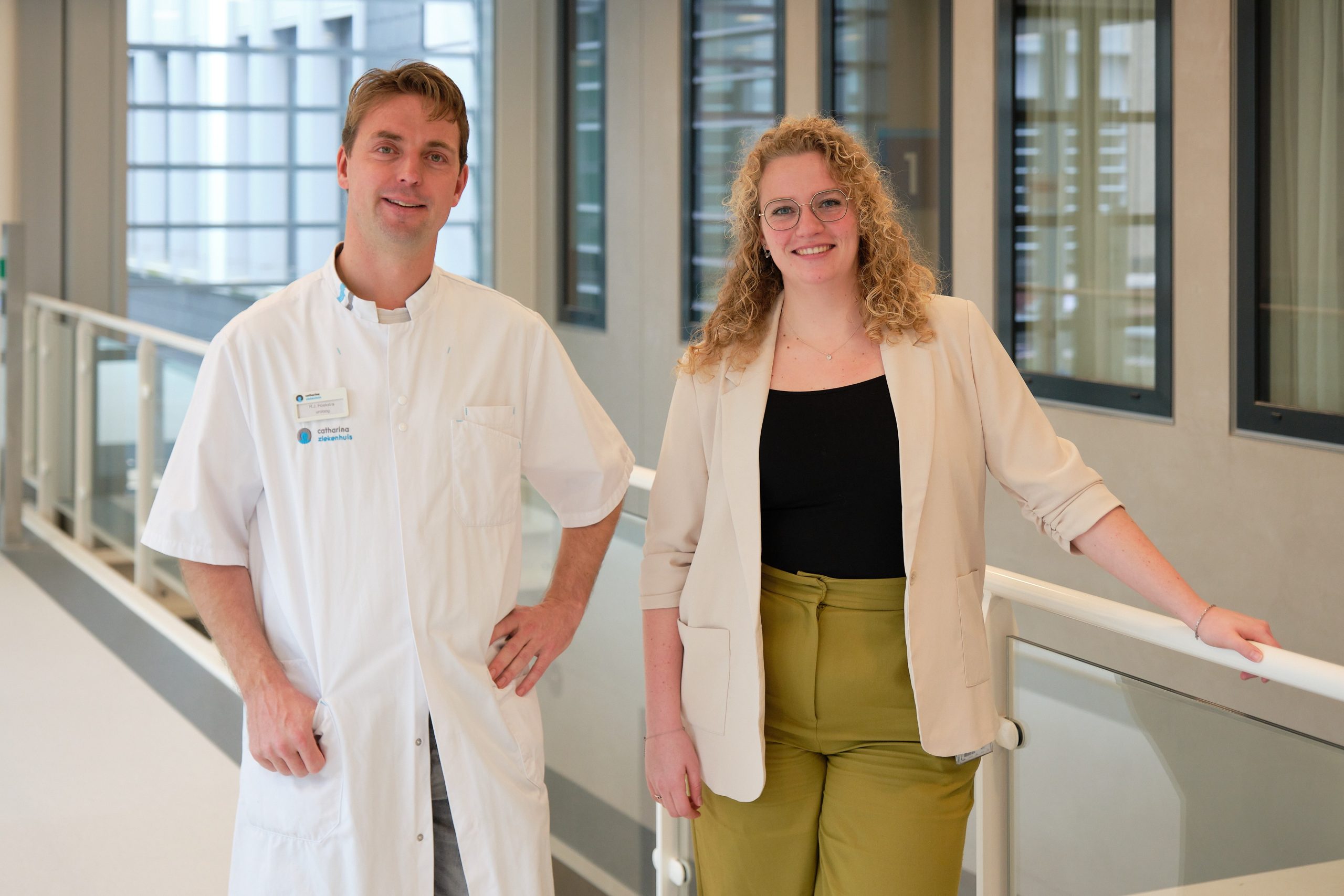 Een man in een witte doktersjas en een vrouw in zakelijke kleding staan lachend naast elkaar in een lichte bende met grote ramen.