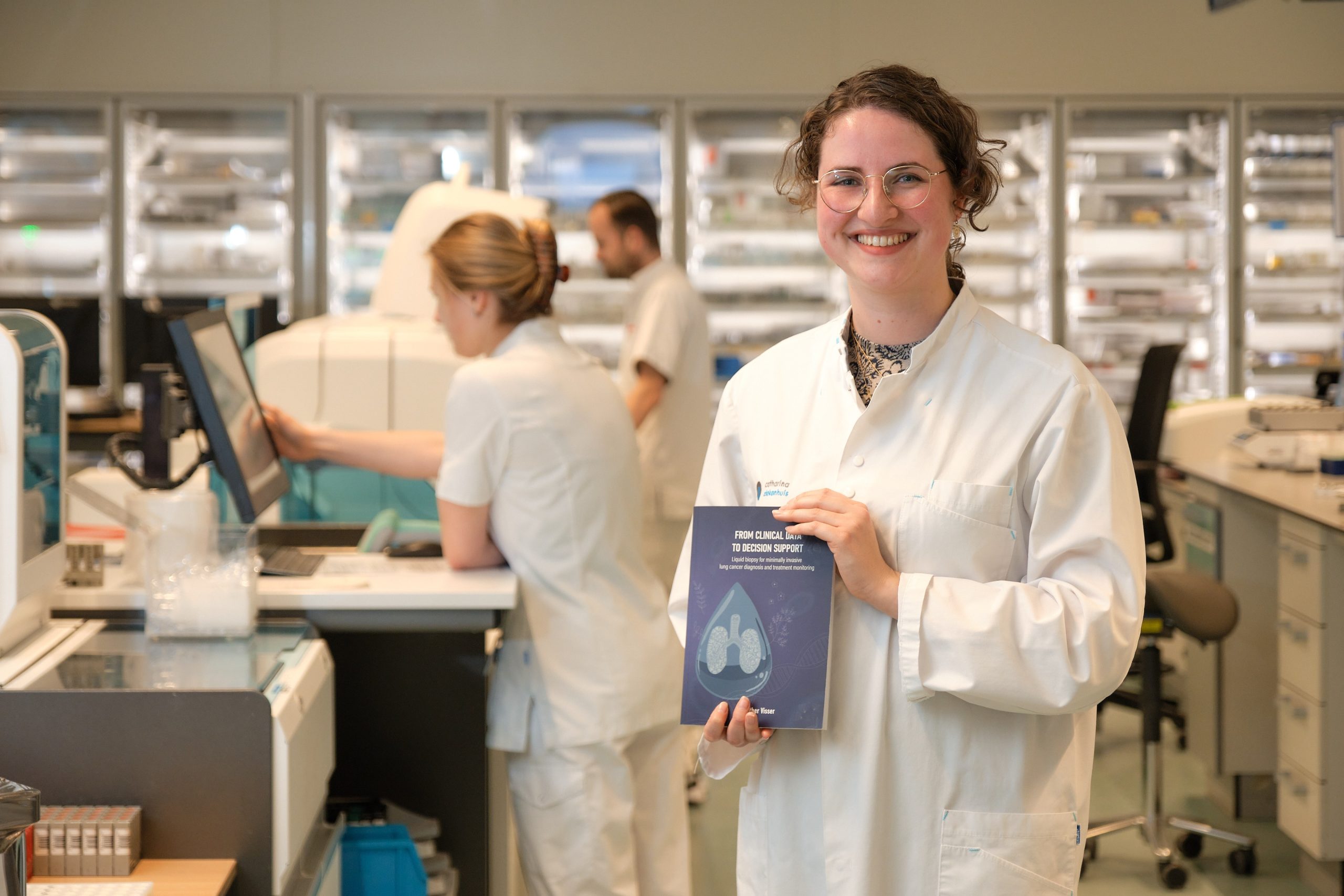 Persoon in witte labjas houdt een boekje met longafbeelding vast; twee anderen werken op de achtergrond in het laboratorium.