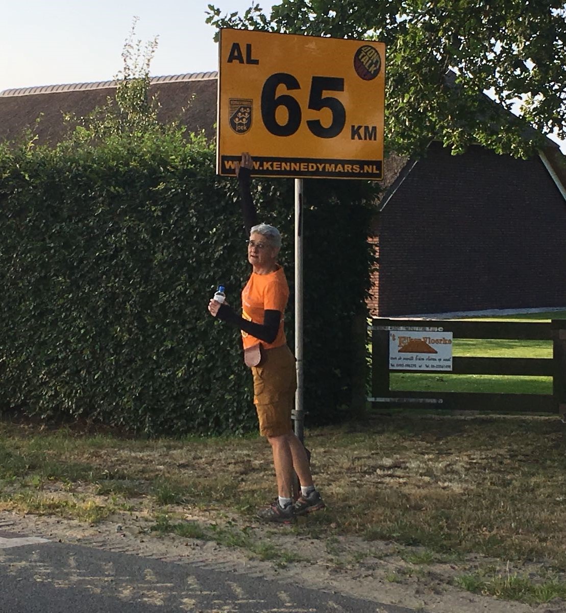 Persoon in oranje shirt en bruine broek raakt tijdens Kennedymars gele 65 KM-bord aan, met heg, gras en gebouwen op achtergrond.