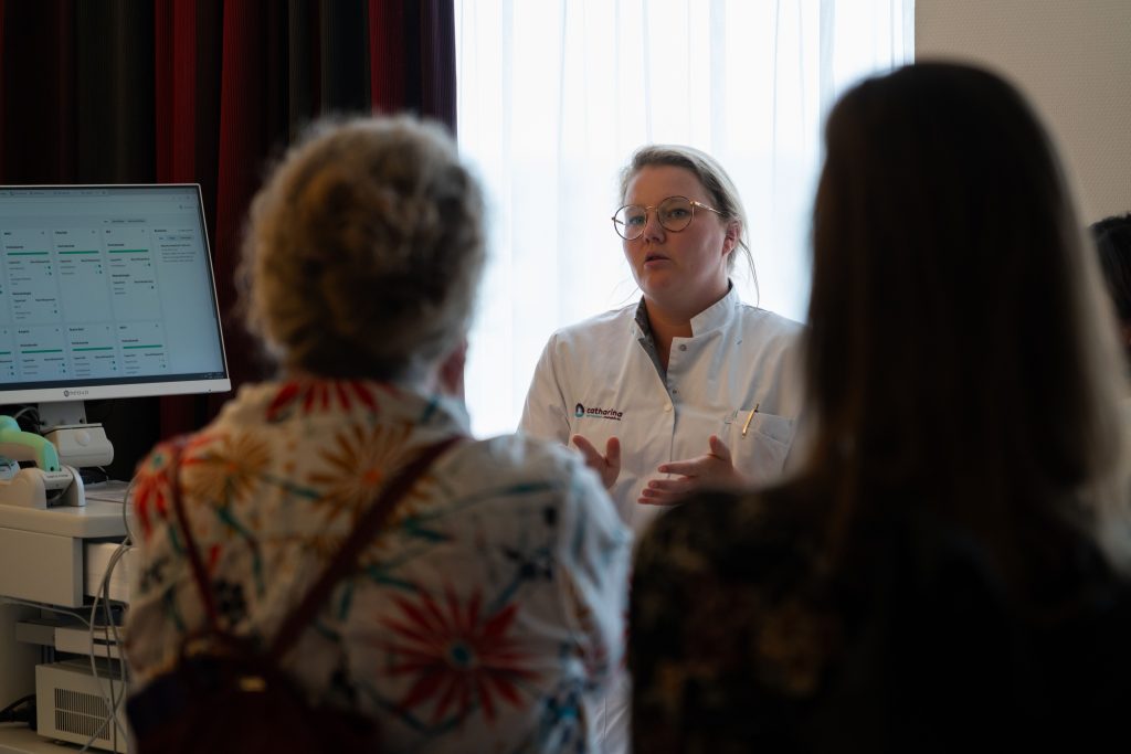 Een zorgverlener in witte jas spreekt met twee collega's in een ziekenhuisruimte met raam en computerscherm met data.