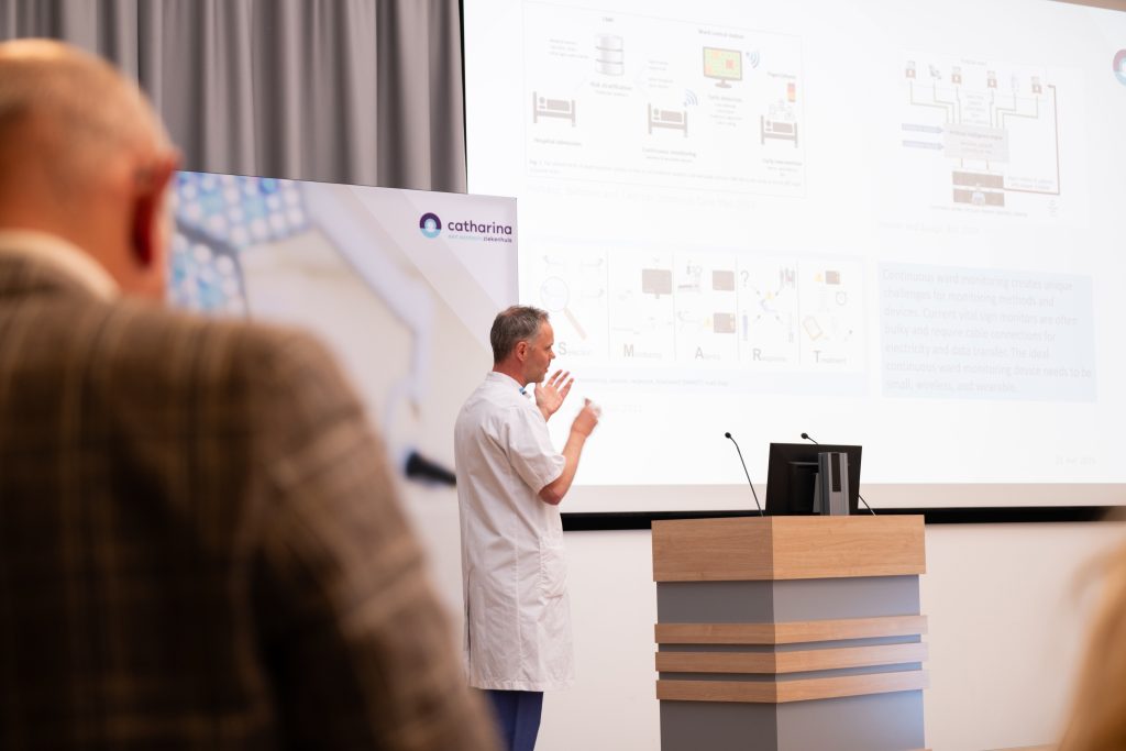 Een man in een witte labjas geeft een presentatie met diagrammen, terwijl iemand in ruitjas toekijkt op Experience Day.