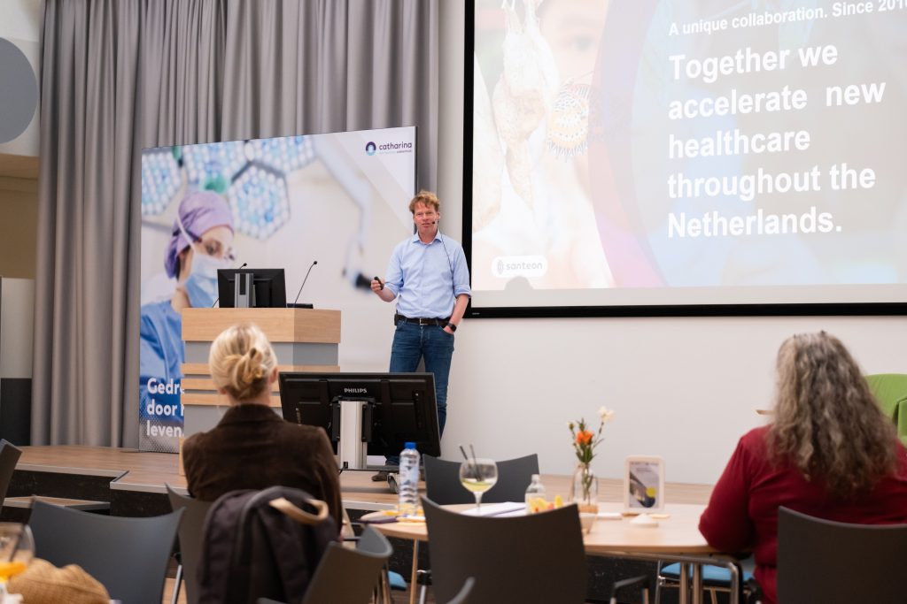 Een man spreekt op een podium naast een presentatie over Nederlandse zorg, buitenlandse collega's zitten aan tafels op de voorgrond.