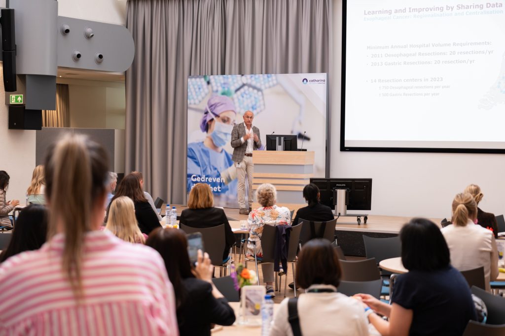 Man geeft presentatie op podium; publiek, waaronder buitenlandse collega's, luistert en maakt aantekeningen; dia's over volume-eisen.