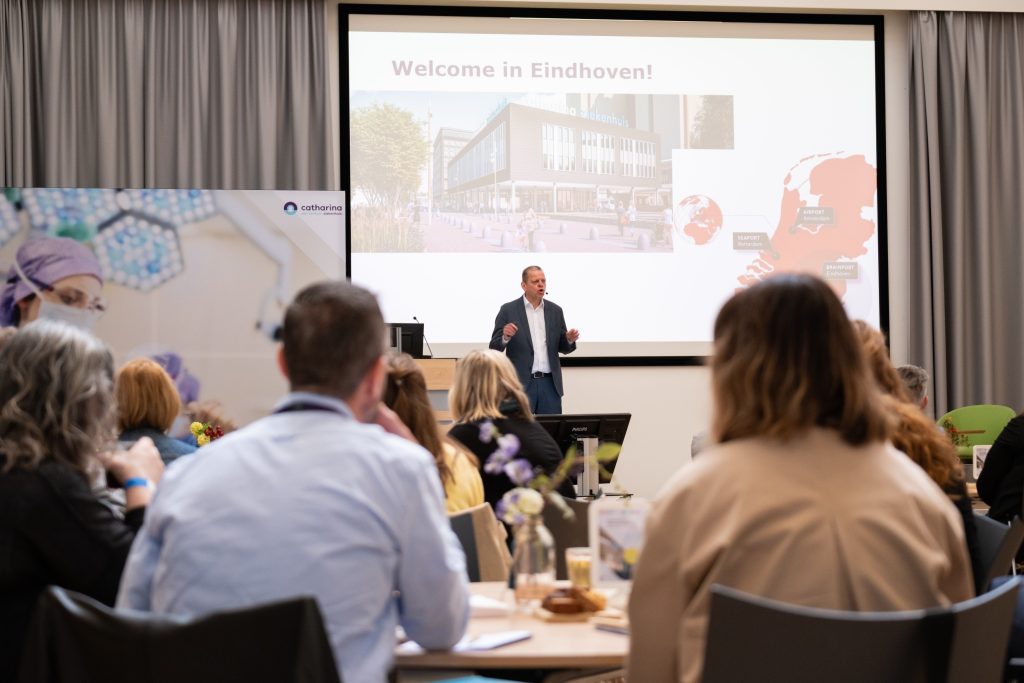 Man in pak geeft presentatie aan buitenlandse collega's; scherm toont "Welcome in Eindhoven" met ziekenhuis en kaart.