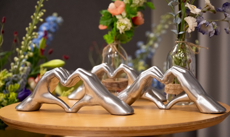 Drie metalen handbeelden in hartvorm op een ronde houten tafel, met glazen vazen en bloemen op de achtergrond.