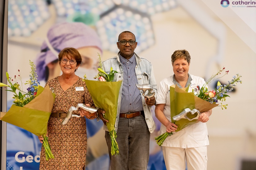 Drie mensen binnen, elk met een bloemenboeket en zilveren prijs. Op de achtergrond logo en iemand in medische kleding, 2025 zorgtoppers.