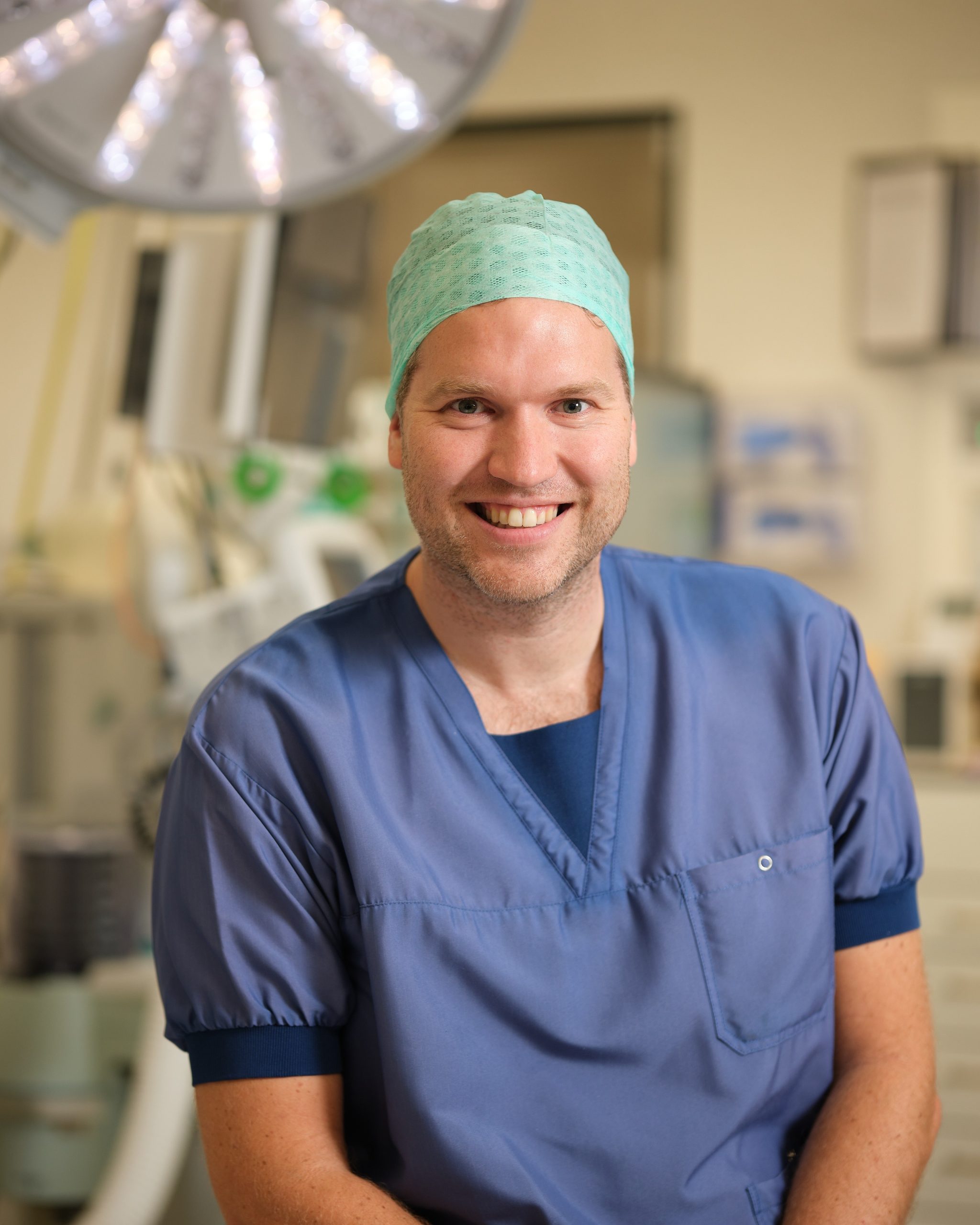 Iemand met blauwe schrobkleding en groene operatiemuts zit lachend in een heldere kliniek, bestralingapparatuur op de achtergrond.