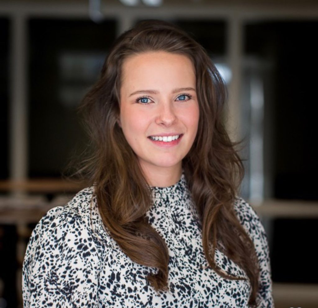 Vrouw met lang bruin haar in witte blouse met zwarte stippen glimlacht binnen; vage ramen en donkere meubels op achtergrond.