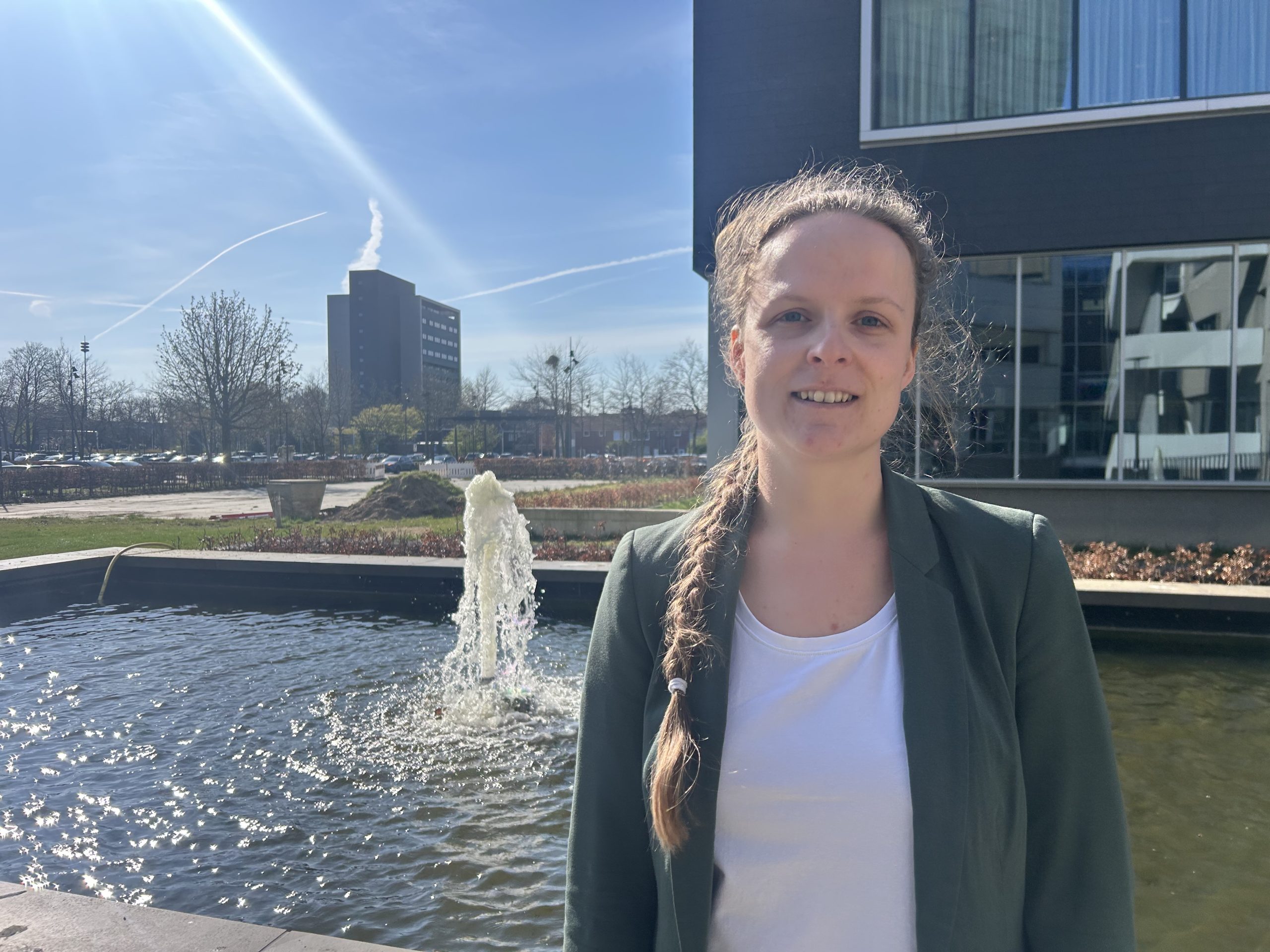 Vrouw met lange vlecht in groene blazer en witte blouse voor fontein bij modern gebouw op zonnige dag, bomen op achtergrond.