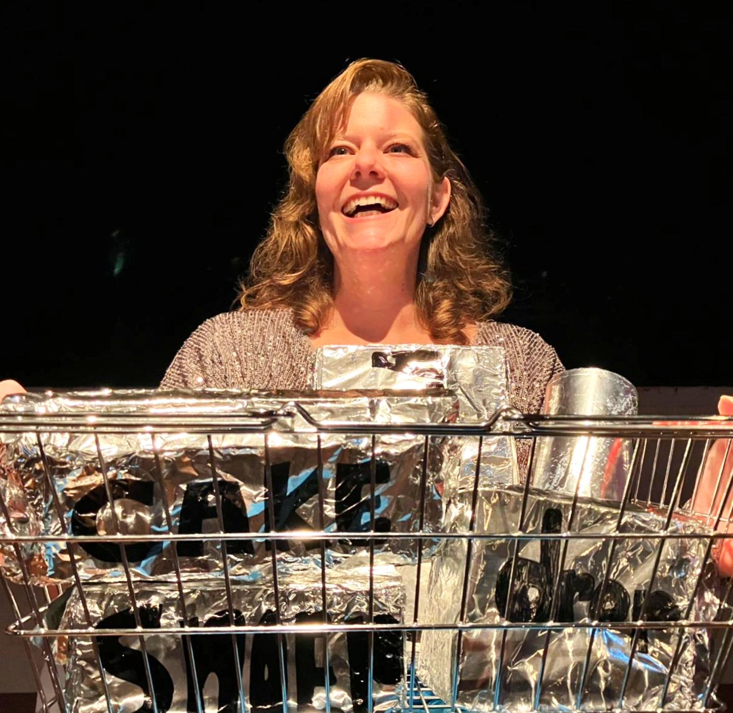 Vrouw met golvend bruin haar glimlacht achter een winkelwagen vol zilverkleurige verpakkingen en flessen tegen donkere achtergrond.