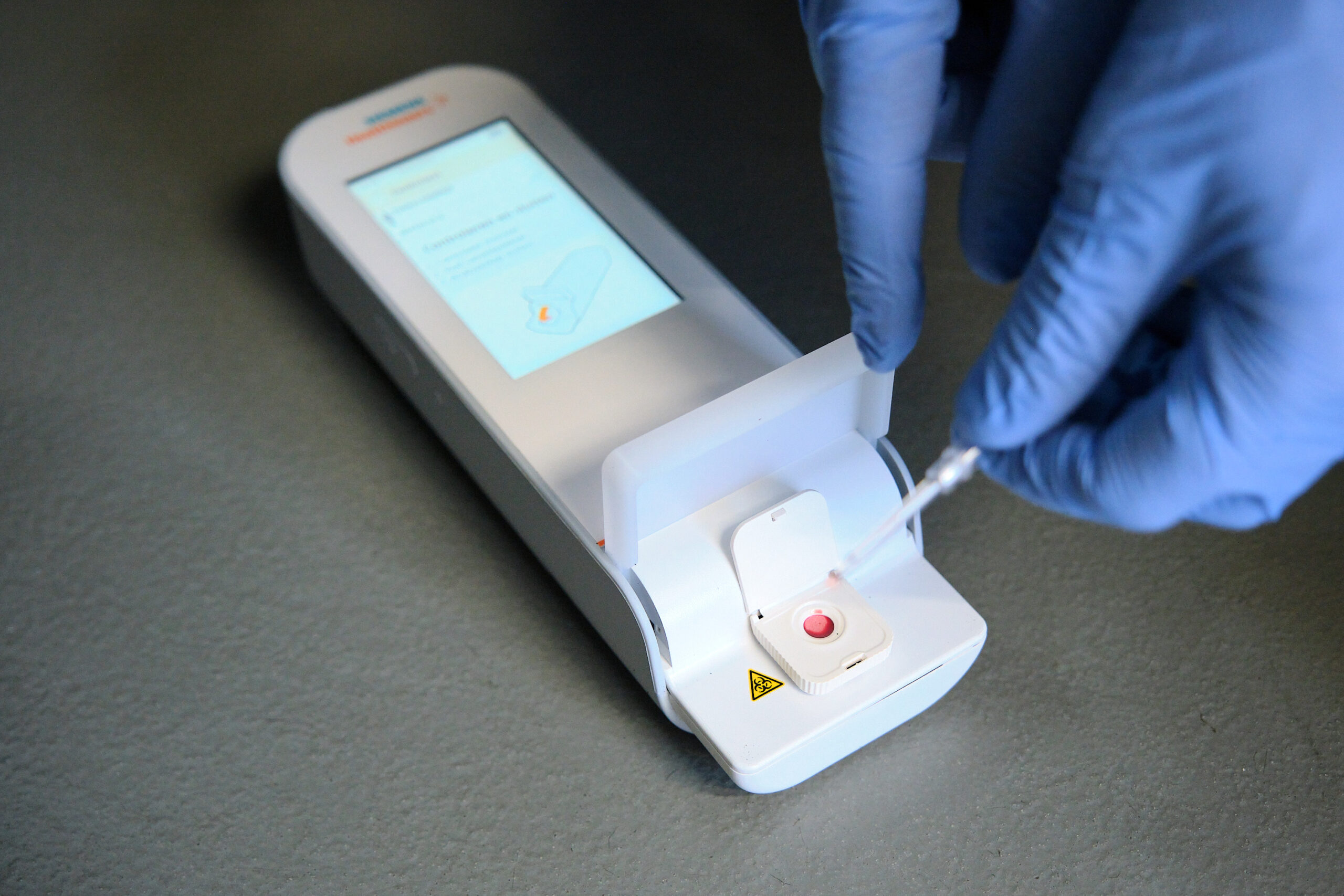 Een gehandschoende hand brengt een pipet met bloed aan op een sneltestapparaat met scherm en compartiment in de Brainportregio.