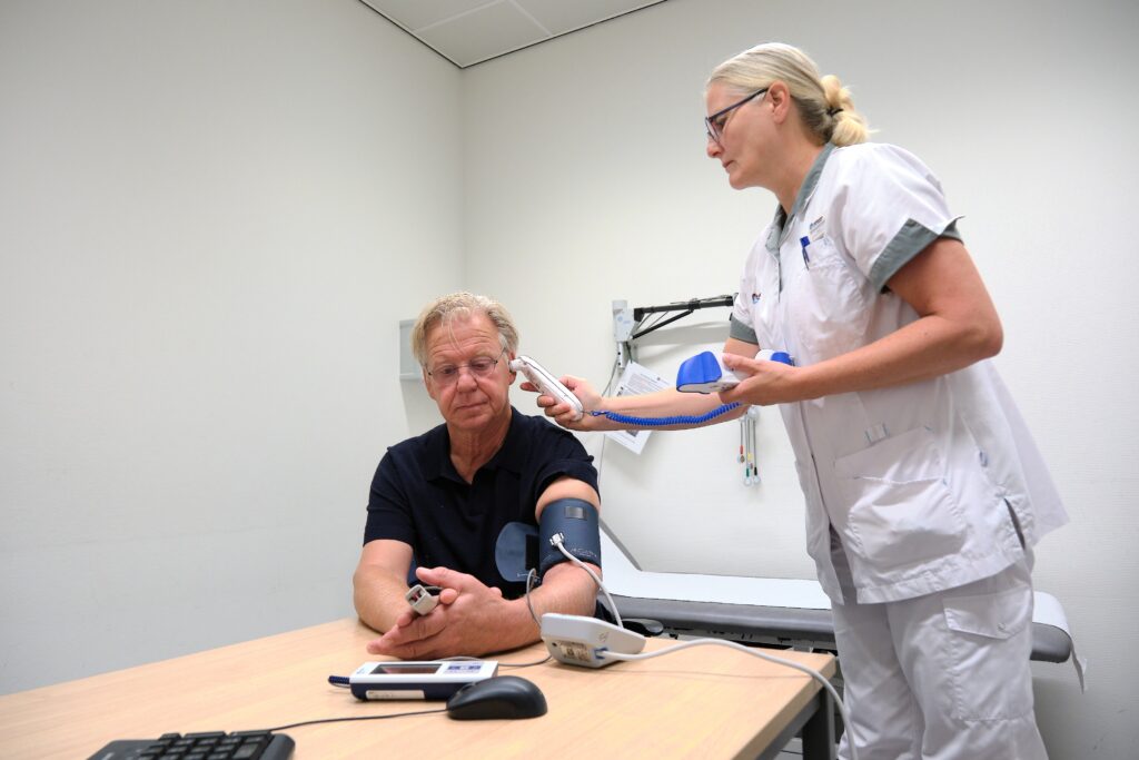Een verpleegkundige ontmoet de bloeddruk van een zittende man met een elektronische meter; andere patiënten worden voorbereid.