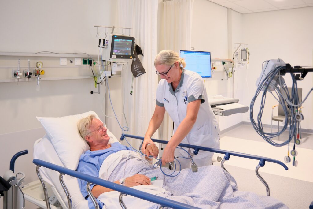 Een verpleegkundige stelt apparatuur af bij een patiënt in bed. De patiënt glimlacht. Medische apparatuur staat op de achtergrond.
