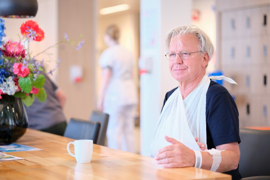 Oudere man met bril en mitella zit aan tafel met mok en bloemen; draagt ziekenhuisbandje. Op de achtergrond zorgpersoneel.