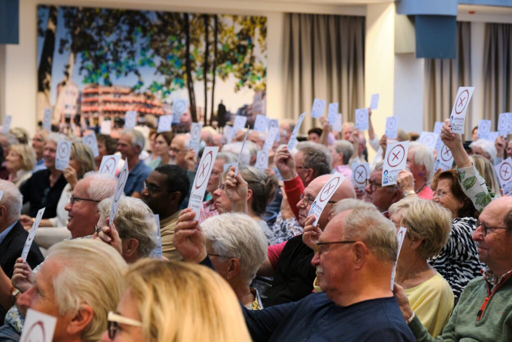 Grote groep ouderen zit dicht bij elkaar in een zaal en houdt kaart met X omhoog. Op achterwand hangt foto van een stad.